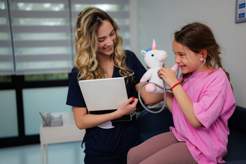 Smiling female pediatrician showing digital tablet to young girl playing doctor with plush unicorn toy and stethoscope in bright modern clinic
