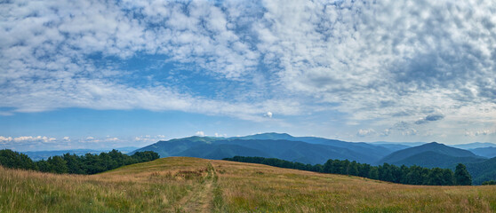 A narrow trail leads over a golden grassy hilltop under a dramatic, cloudy sky, with panoramic views of rolling hills and distant valleys stretching to the horizon.