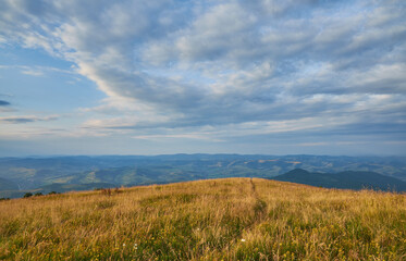 A narrow trail leads over a golden grassy hilltop under a dramatic, cloudy sky, with panoramic...