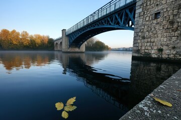 Fototapeta premium Calm waters reflect an autumn bridge in early morning light near a peaceful riverbank
