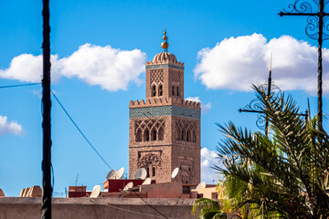 Koutoubia Mosque minaret rising above rooftops and palm trees in Marrakesh, Morocco, under a blue sky, showcasing historic Islamic architecture, cultural heritage and the vibrant cityscape atmosphere.