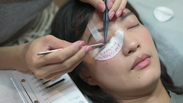 Southeast Asian woman receiving eyelash extension treatment beauty salon, eyes closed and relaxed as technician applies individual lashes with tweezers, showcasing eye and face beauty enhancement
