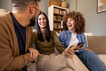 Young Diverse Friends Share Joyful Moment On Living Room Couch With Mobile Phones And Natural...