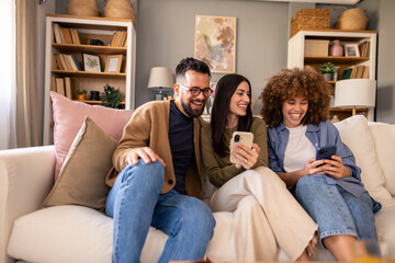 Diverse Group Of Friends Lounging On White Couch Using Smartphones Together In Cozy Home Library...