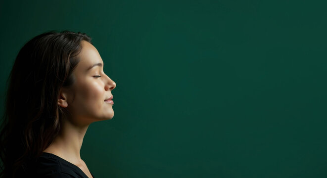 Side profile of woman with dark hair on deep green background. Serene expression with closed eyes and black top. Mindfulness and inner calm. Meditation programs, yoga classes, mental wellness, stress
