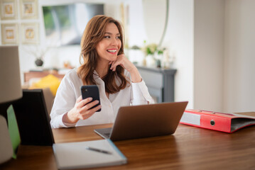 Obraz premium Confident woman sitting in front of her laptop and using her smart phone while working from home