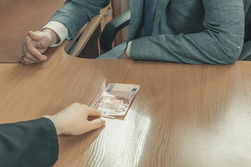 Hand moves euro bills across an office table towards a man in a suit and tie sitting opposite. Give a bribe. Corruption