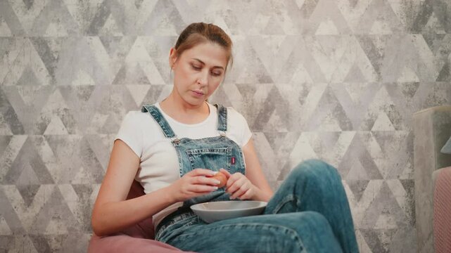 Portrait view of relaxed woman in denim overalls seated on bean bag cracking boiled egg on glass plate, with soft lighting and geometric wallpaper in background