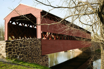 A rustic red covered bridge over a calm river, framed by stone walls and bare trees. Perfect for themes of history, architecture, or tranquil landscapes.