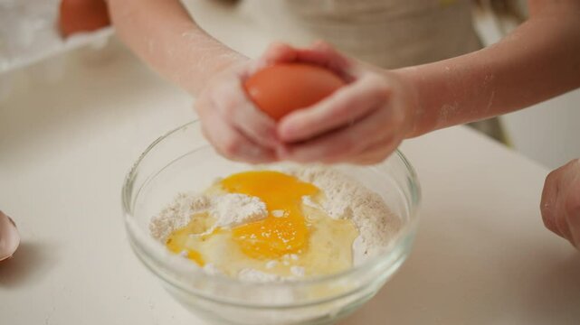 hand view of person breaking brown egg into bowl filled with flour and cracked yolk, preparing ingredients for baking on white surface, showcasing hands-on kitchen