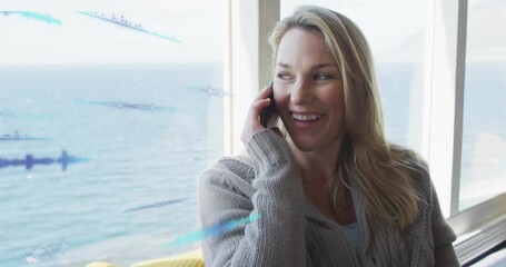 woman sitting by window facing sea, talking on smartphone, showing technology waveform overlay - Powered by Adobe