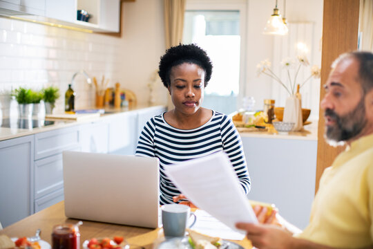 Worried couple reviewing financial documents together at kitchen table with laptop open