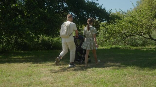 Young parents spend time together walking with their child in the park. Mom and dad push a stroller with their baby across the grass, seeking shade under the trees on a hot sunny day.