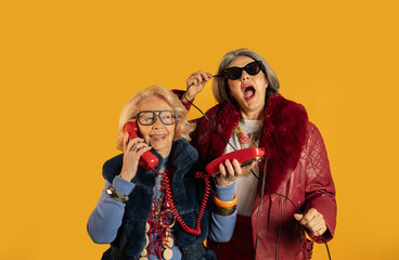 Two senior women having fun with vintage phones on yellow background