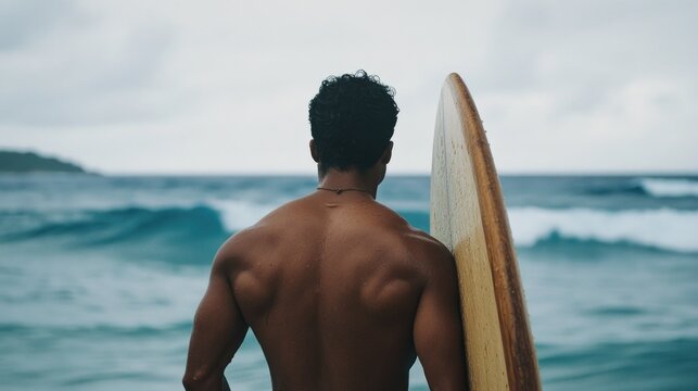 Young male surfer with surfboard facing ocean waves on cloudy day