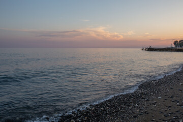 Obraz premium A calm twilight settles over the quiet Mediterranean shoreline. Gentle waves lap the sand, and pastel hues fill the sky as evening falls on the peaceful, nearly deserted beach.