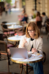 Mid aged businesswoman sitting at outdoor cafe and working on her laptop