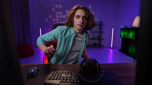 Young adult man spending time at home, man streamer gamer sits at desk starts recording stream live, talking at camera on computer, playing game in headphones, person pov.