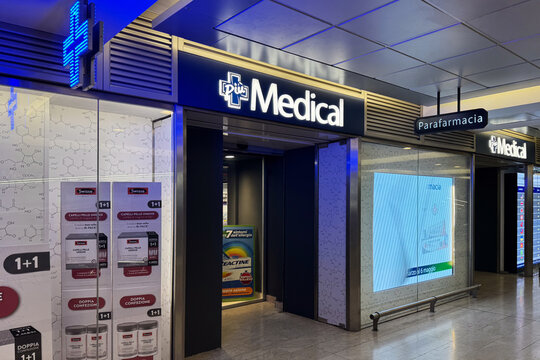 Milan, Italy - April, 07, 2025: Piu Medical Parafarmacia drugstore with glowing pharmacy cross and health posters inside Milan central train station.
