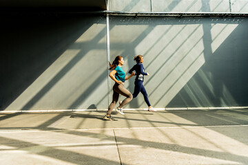 Two women jog energetically under warm sunlight with striking shadows creating dynamic patterns on...