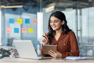 Fototapeta premium A businesswoman smiles while working on her laptop and taking notes in an office environment. Sticky notes are on the window behind her.
