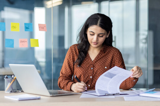 A focused woman, working intently on paperwork at a desk in a modern office setting. A laptop sits beside her, and sticky notes are visible.