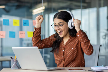 A joyful woman celebrates a win, wearing headphones and glasses, looking at her laptop in an office...