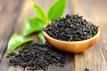 Dried tea leaves in a wooden bowl on rustic surface. Fresh green leaves nearby suggest natural origin and quality of the product.