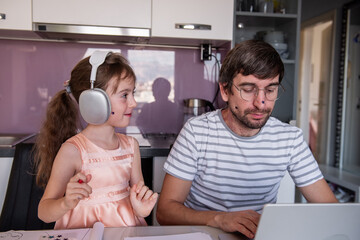 Father working remotely at the kitchen table while daughter plays and enjoys music through...