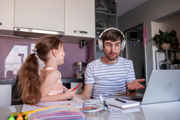 Father wearing headphones is working from home, having a video conference on his laptop while taking care of his little daughter who is drawing next to him in their kitchen