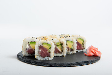 Close-up of sushi California rolls filled with tuna, avocado, and sesame seeds served on a black slate plate, accompanied by pickled ginger on isolated white background