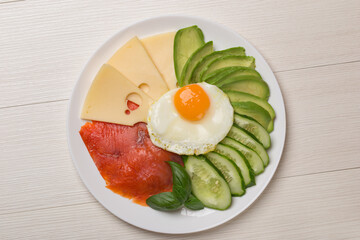Top view on plate with scrambled eggs, salmon, cheese, cucumbers and avocado on a light wooden. Healthy eating plate.