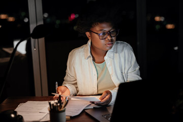 Young African businesswoman working on a laptop and paperwork in the evening