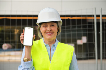 Smiling female engineer wearing safety helmet and vest showing smartphone with blank white screen...