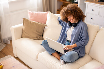 Obraz premium Casual Remote Worker With Natural Curly Hair Using Laptop On Cozy White Sofa In Bright Home Interior