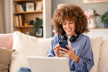 Curly-Haired Woman Smiling Indoors with Laptop Headphones and Mobile Phone on a Cozy Couch