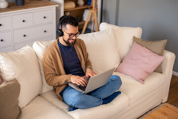 Fototapeta premium Indoor Caucasian Man Sitting on Couch Wearing Headphones and Using Laptop in Bright Living Room