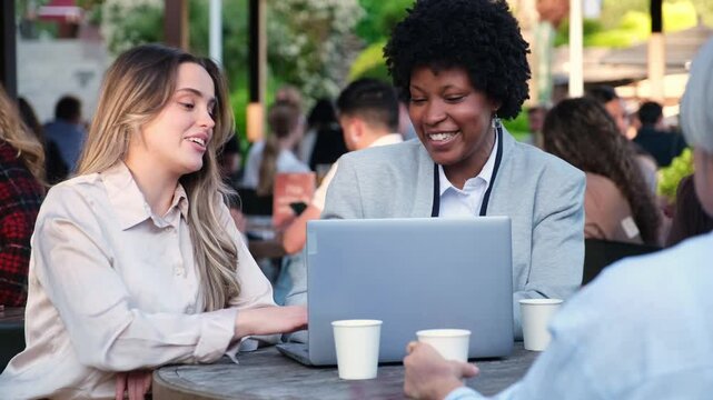Two businesswomen are discussing a project on a laptop in a crowded outdoor cafe
