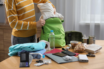 Survival kit. Woman packing different emergency supplies into backpack at wooden table indoors,...