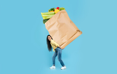A customer joyfully carries a heavy paper bag filled with fresh vegetables and fruits in a store....