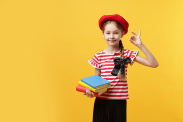 Schoolgirl with beret and books showing okay gesture on yellow background. Space for text