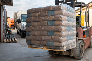 A forklift moves a pallet of plastic wrapped bags in an industrial area. A white van, orange cement mixer, and scattered wooden pallets are visible.