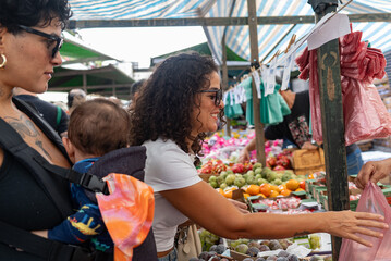 Casual street market scene of two Brazilian women and baby shopping for vegetables

