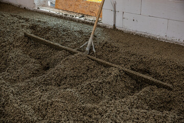 An indoor construction site shows wet concrete being leveled with a rake like tool. Unfinished...