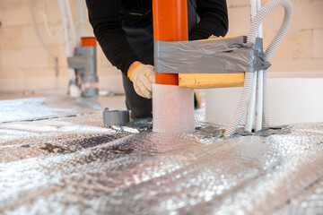 A worker secures red underfloor heating pipes with duct tape and foam insulation on reflective foil. Tools, materials, and unfinished walls are visible.