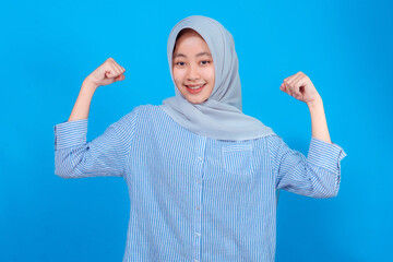 Cheerful young Muslim woman in hijab smiling brightly while showing both flexed arms, symbolizing...