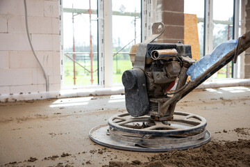 Interior of a construction site showing a power trowel machine on a concrete floor, unfinished walls, large windows, and visible underfloor heating pipes.