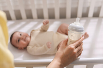 Mother with feeding bottle of milk near her little baby indoors, selective focus
