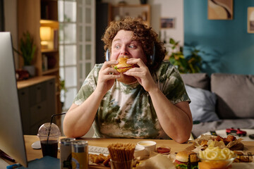 Person with curly hair enjoying a large sandwich in a cozy, cluttered room filled with various...