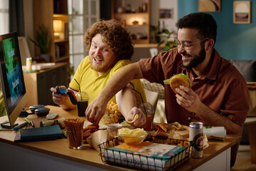 Two friends enjoying snacks and drinks while focusing intently on video games. Modern apartment setting with various snacks and beverages spread out on the table